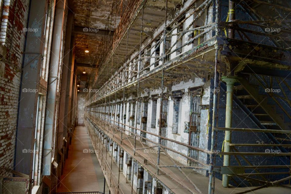 A long hallway of jail cells with rusty metal bars and narrow walkways with multiple tiers 