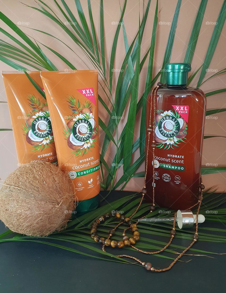 Photo of XXL Herbal Essences Coconut Scent shampoo and conditioner placed on black paper and palm leaves with a brown paper background together with a coconut, dropper and brown beads necklace and bracelet