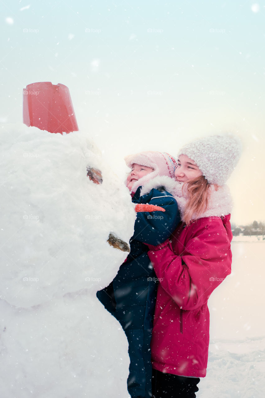 Girl and her little sister making a snowman, spending time together outdoors, having fun in wintertime