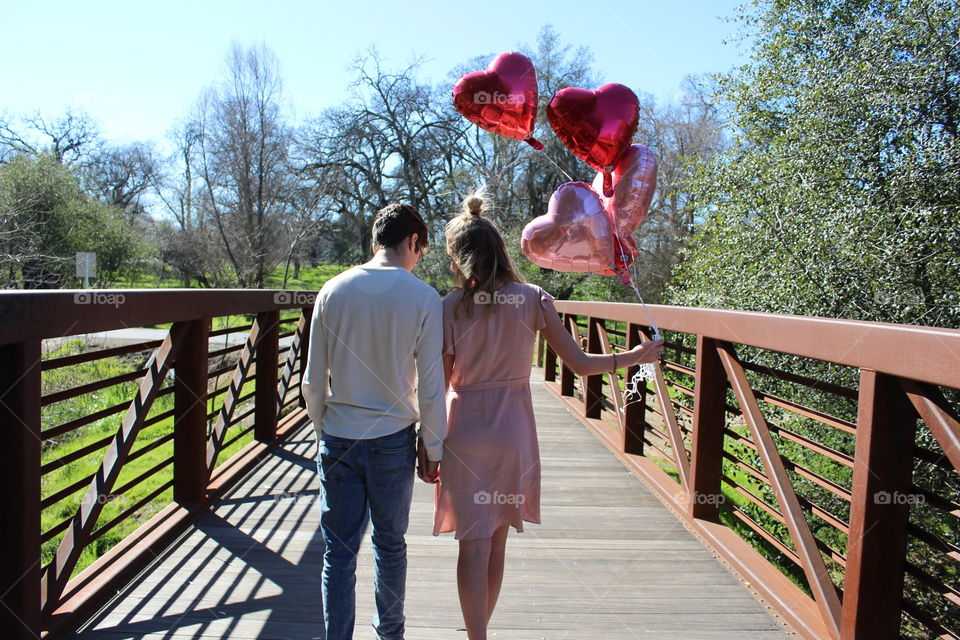 A boy and girl holding hands while walking over the wooden bridge in nature while holding Valentine’s Day pink and red heart shaped balloons. USA, America 