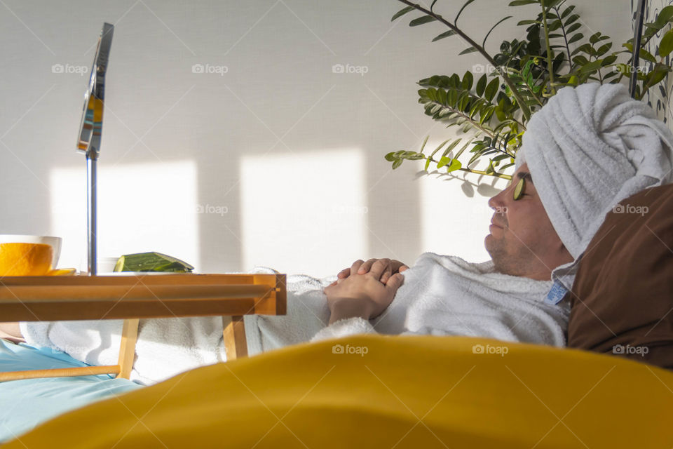 A man and home spa care at home takes care of himself and smears his face with cream, does a manicure at home on a blue background and with oranges and rests.