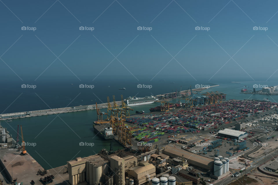 The port of Barcelona seen from above.
View from Montjuic mountain. The sky is clear and the sun is shining. The port is framed by green trees.