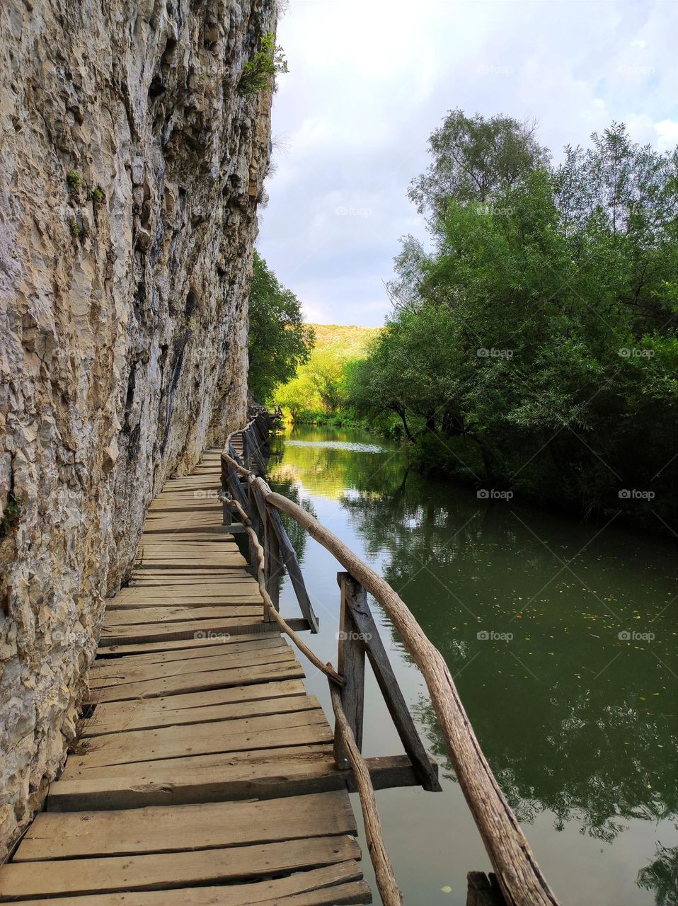 Beautiful wooden pathway on the side of a rock with a calm river under it/ Forest path alongside a very calm and beautiful river with trees on the side of it