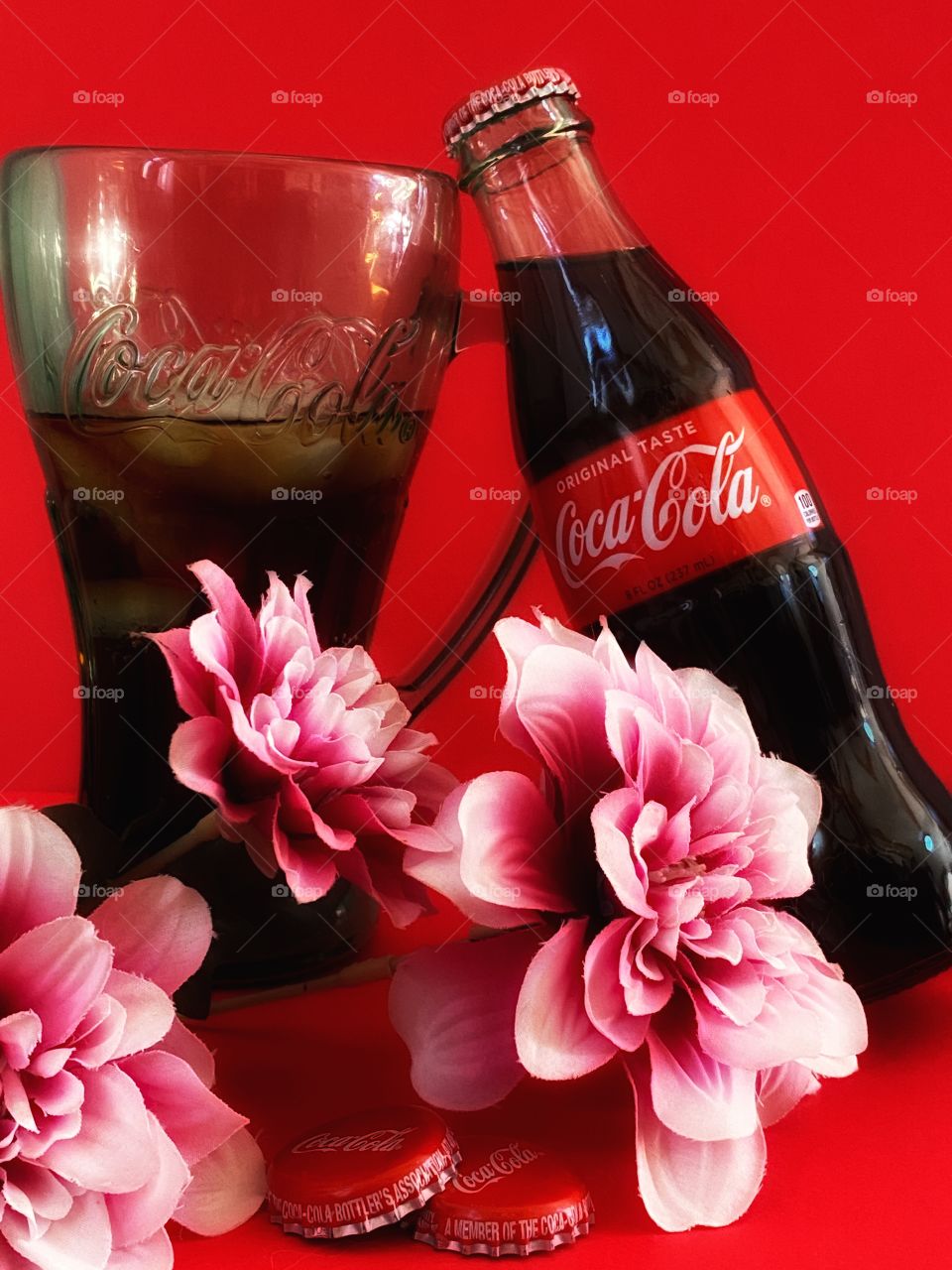A Coca Cola bottle with a Coca Cola glass background is red and tree pink flowers in front 