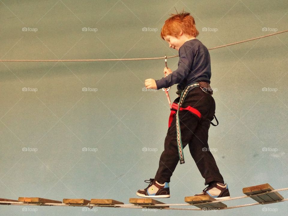 Confident Child On Rope Bridge. Boy In Harness Crossing A Rope Bridge