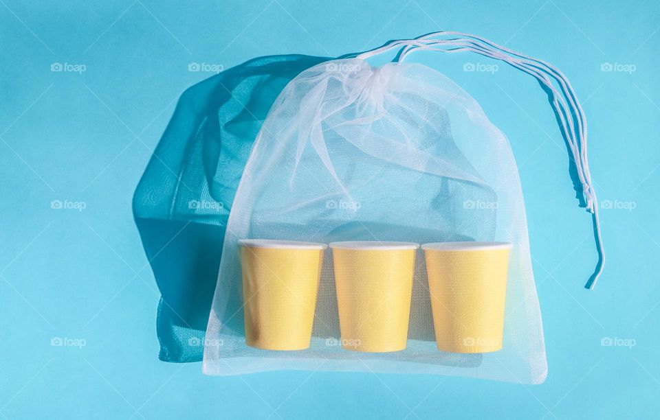 Three yellow cardboard glasses in a transparent mesh bag lie on a blue hair dryer with hard shadows from the sun, flat lay close-up. The concept of paper utensils, ecology day, easter accessories.