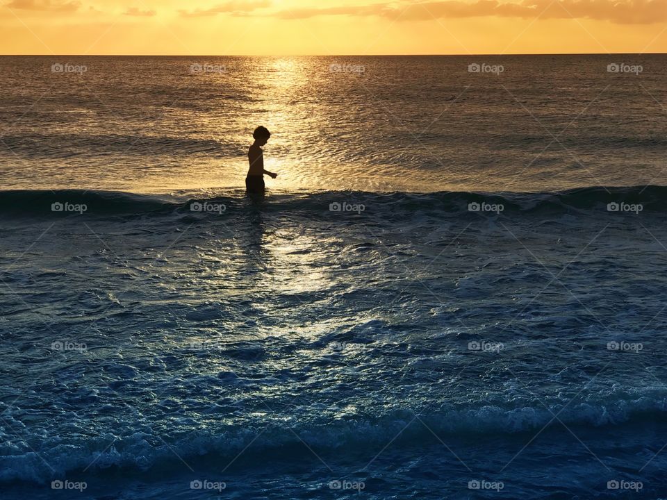 The golden sun reflects on the silhouette of a small boy playing in the ocean.