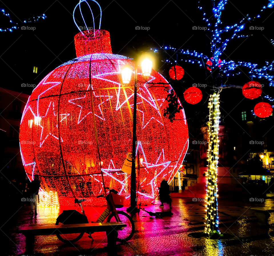 Huge red lit up Christmas bauble and other decorations stand out in the dark of the night, illuminating a bicycle by a bench too. Plaça Da Figueira, Lisbon Christmas 2019