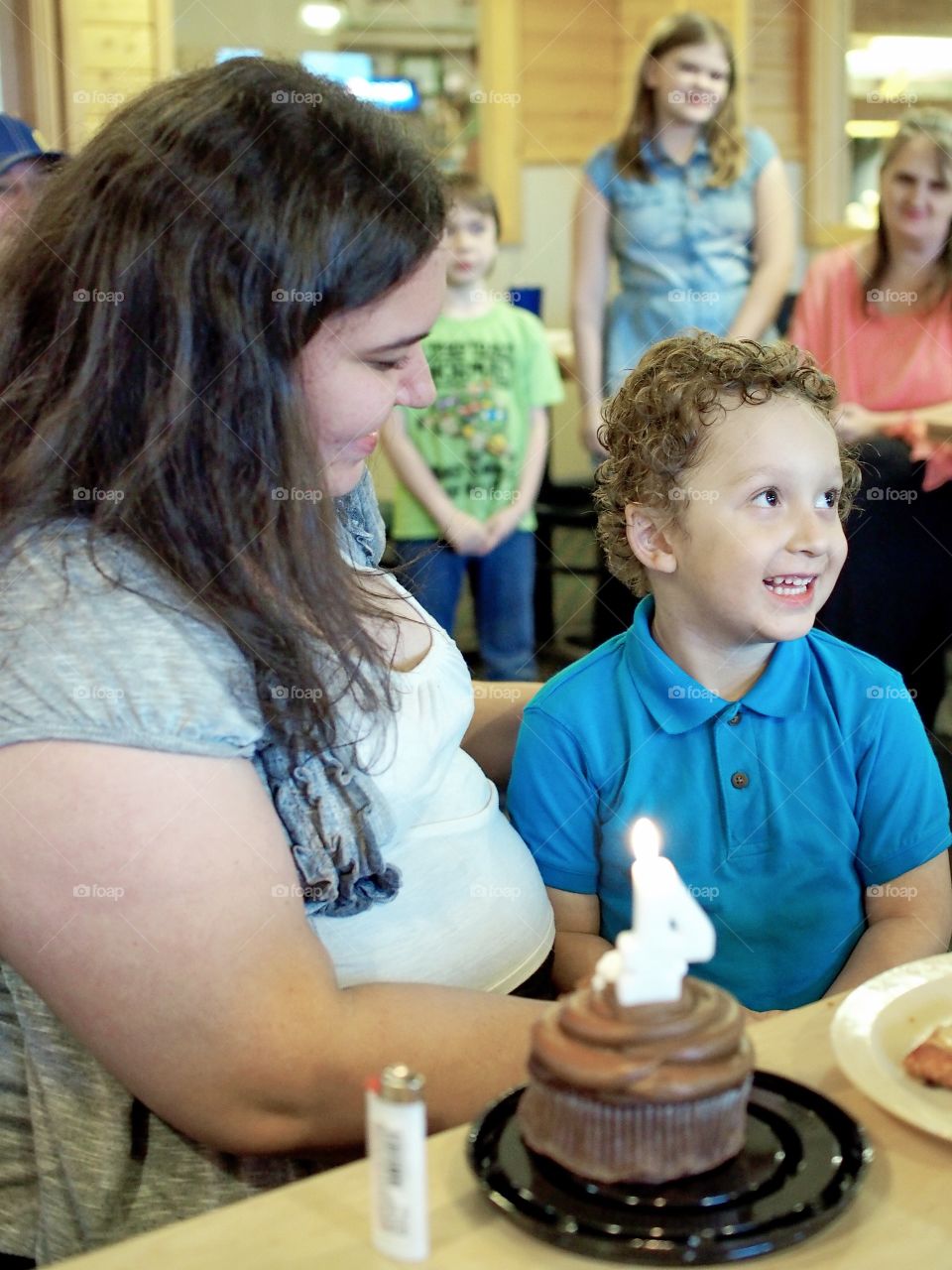A mother holds her delighted son in her lap at his 4th birthday party as the group sings “Happy Birthday” to him. 