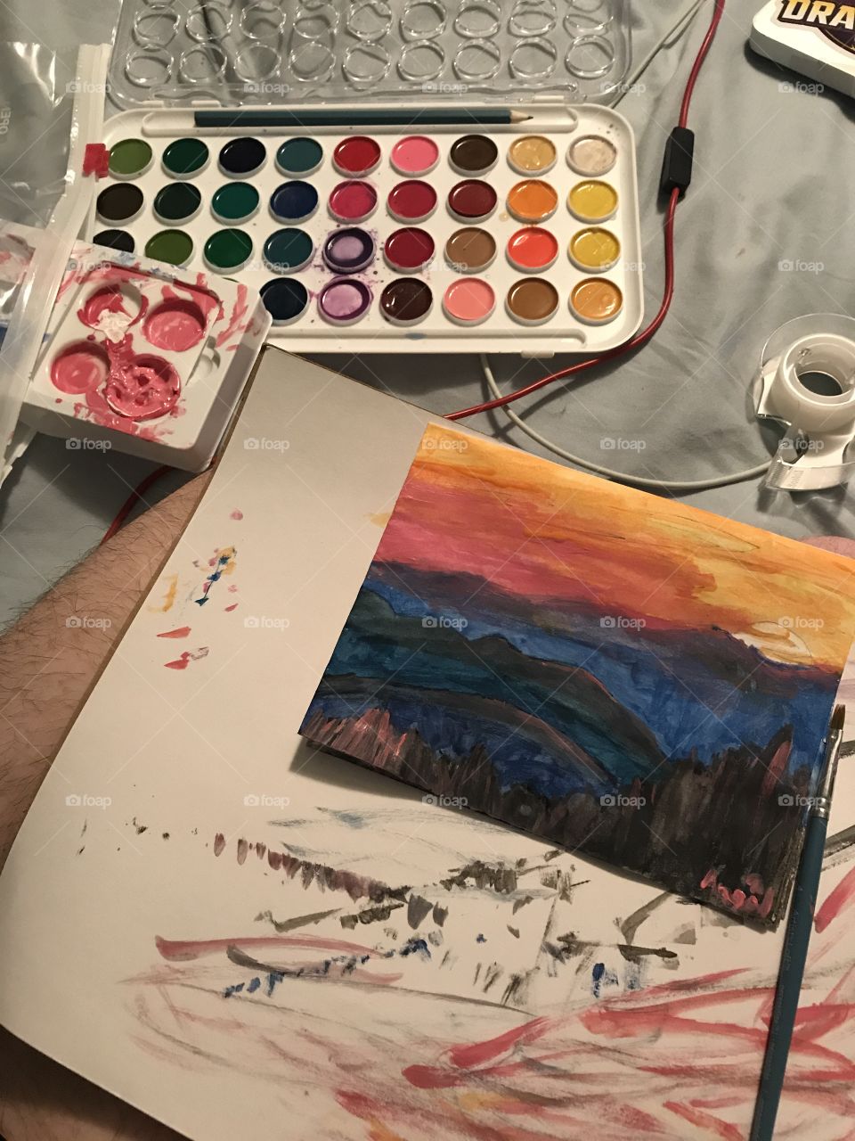 A set of watercolors and acrylics next to a piece of paper, a painting of a sunset in the mountains, and a tape dispenser