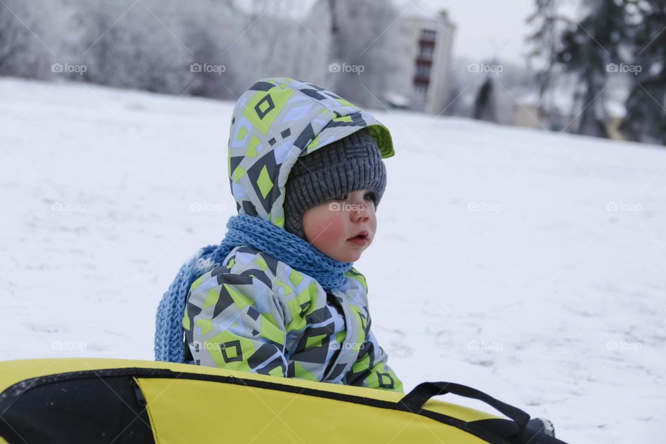 A small, carefree boy walks in the white snow in winter and rides a tubing in the park, near trees in the snow.