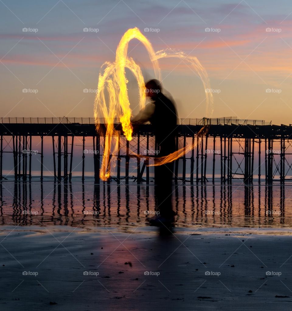 A man spinning fire poi on a beach in front of a pier at dusk