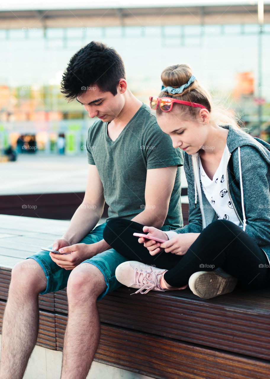 Couple of friends, teenage girl and boy, having fun together, using smartphones, sitting in center of town, spending time together. Real people, authentic situations