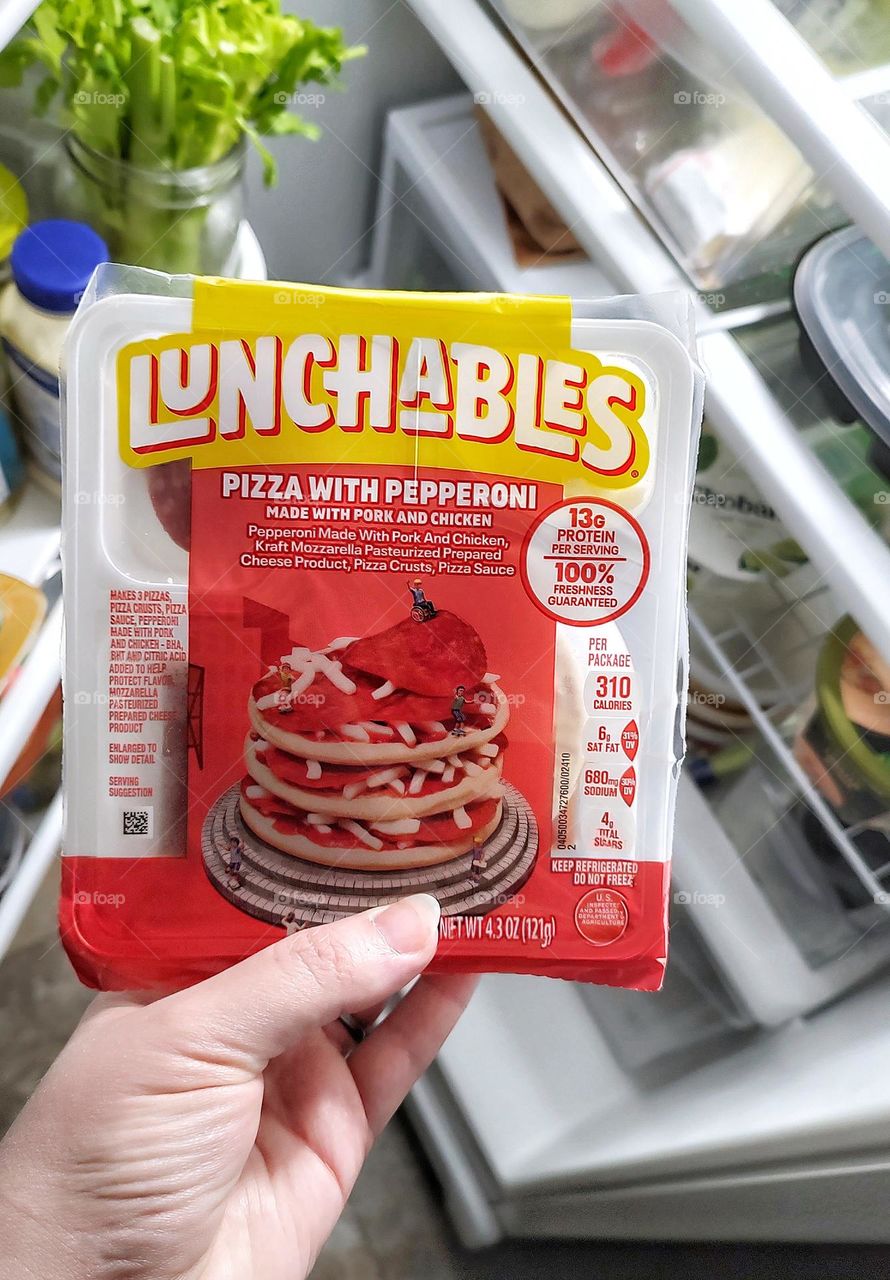 Feminine hand holding up a pepperoni pizza Lunchable in front of an open refrigerator