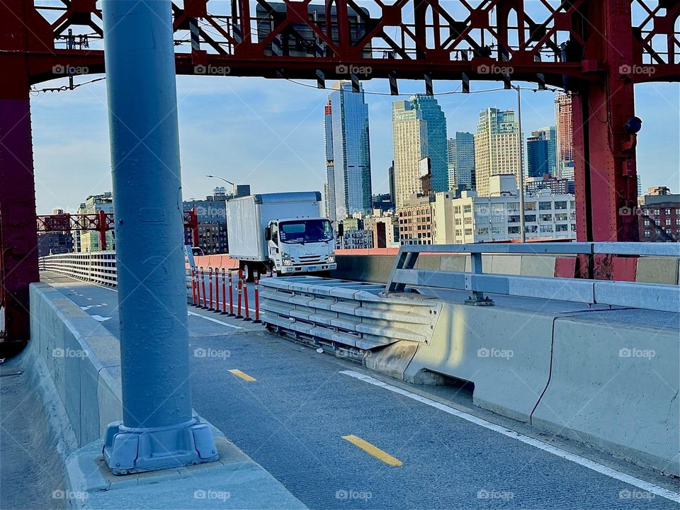 This is the “Pulaski Bridge” at “Newtown Creek” that connects “Greenpoint”, Bklyn to LIC, Queens. Through the ornamental red metal gate we see nearby LIC high rises all aglow in the setting evening sun. 2024. Hypnotic Productions