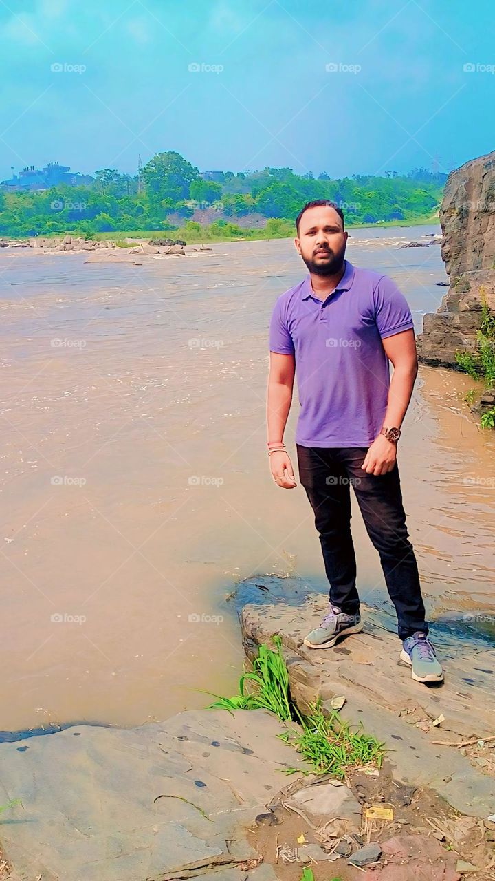 This photograph captures a serene moment in nature, featuring a man standing on the edge of a rocky riverbank.This image embodies themes of peace, harmony, and the simple yet profound connection between humans and the natural world.