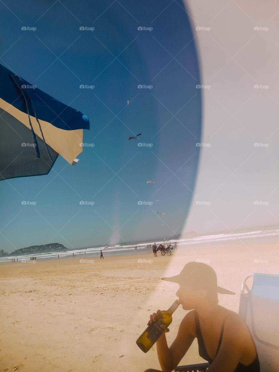Shot of Enseada Beach, Guarujá. There's a girl drinking wine, whit a hat, beneath a shade. In the background we se a kite seller, with his bike.