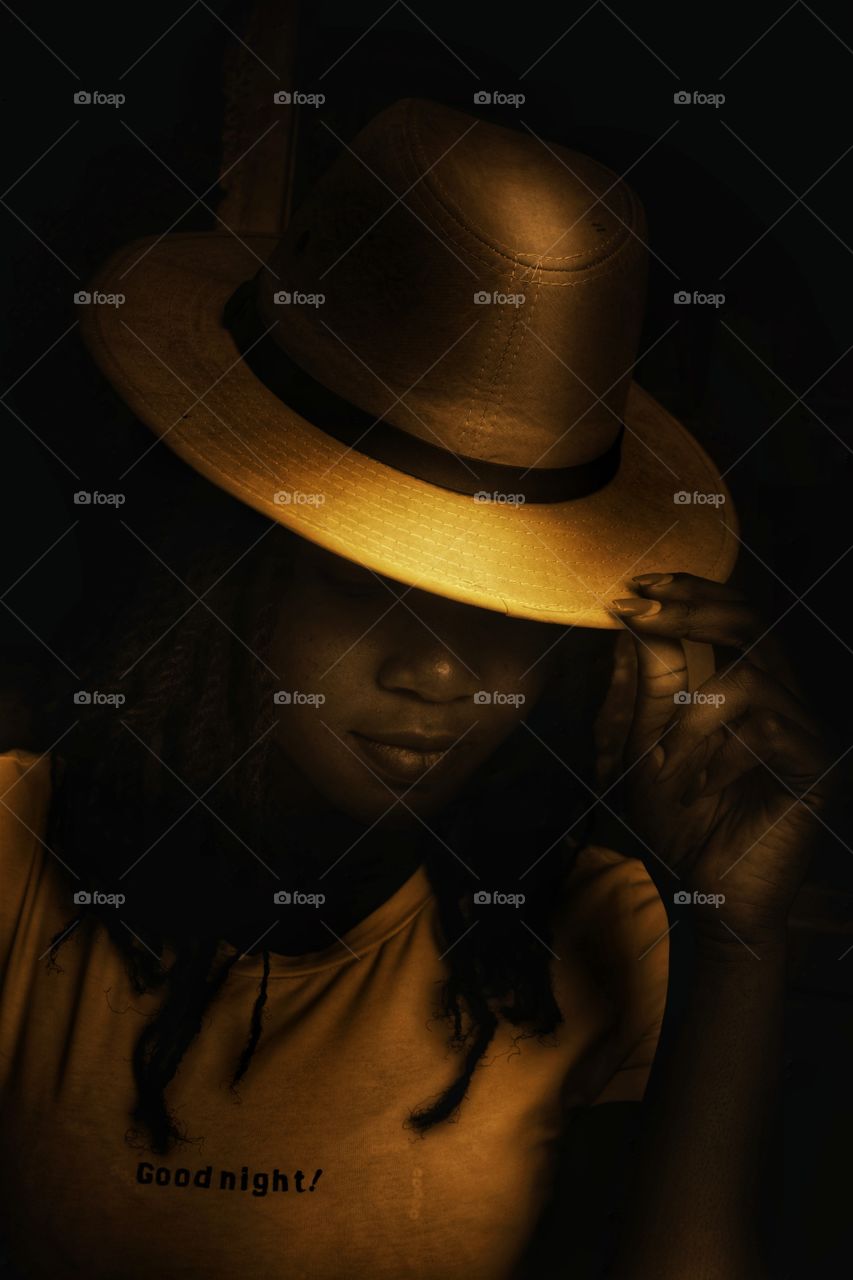 a shadowy portrait of a woman wearing a wide brimmed hat in a dimly lit shadowy room with golden light over her