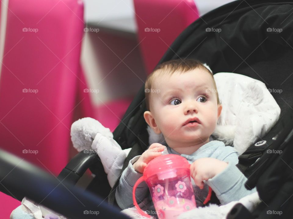 Portrait of one beautiful caucasian baby girl holding a bottle of water in her hands and looking in surprise with curiosity while sitting in a stroller in a restaurant, close-up side view. Surprised people concept.