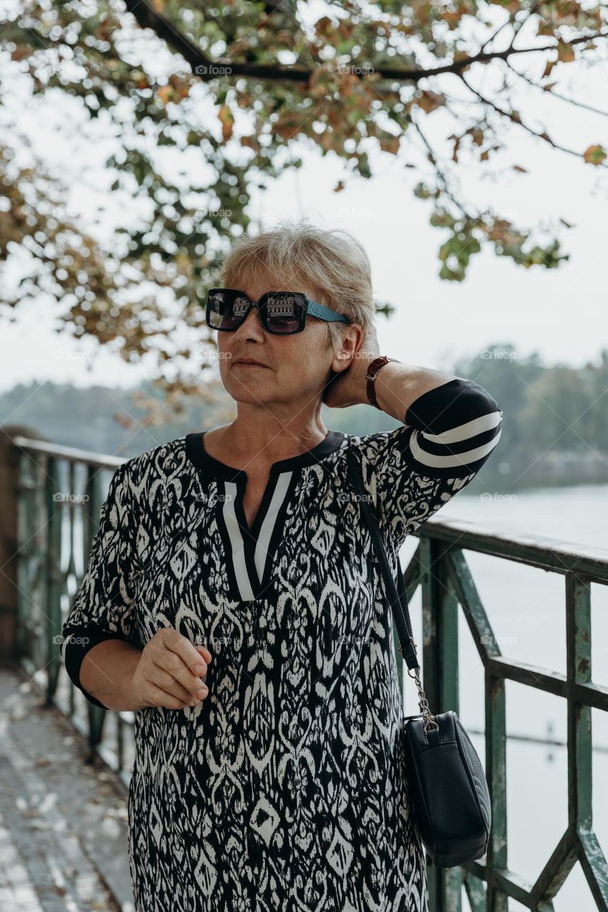 Portrait of a beautiful elderly blonde Caucasian woman wearing a dress and sunglasses, looking away and fixing her hair while walking along a city promenade on an autumn day, close-up side view.