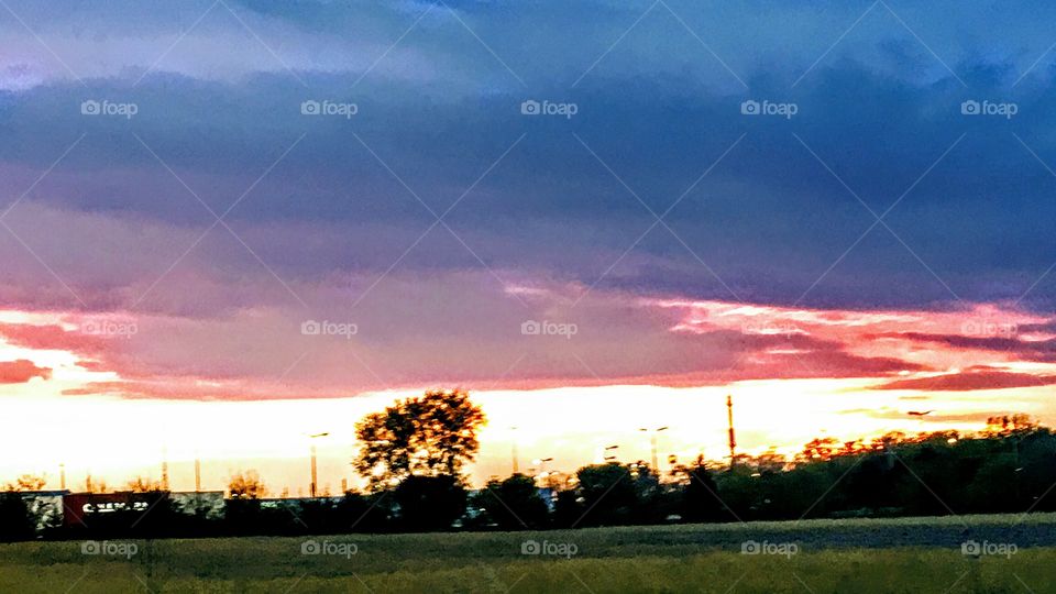 Fiery sunset with deep reds, yellows, oranges, pinks, and purples, through silhouette of trees and commerce, over field in Spring,
