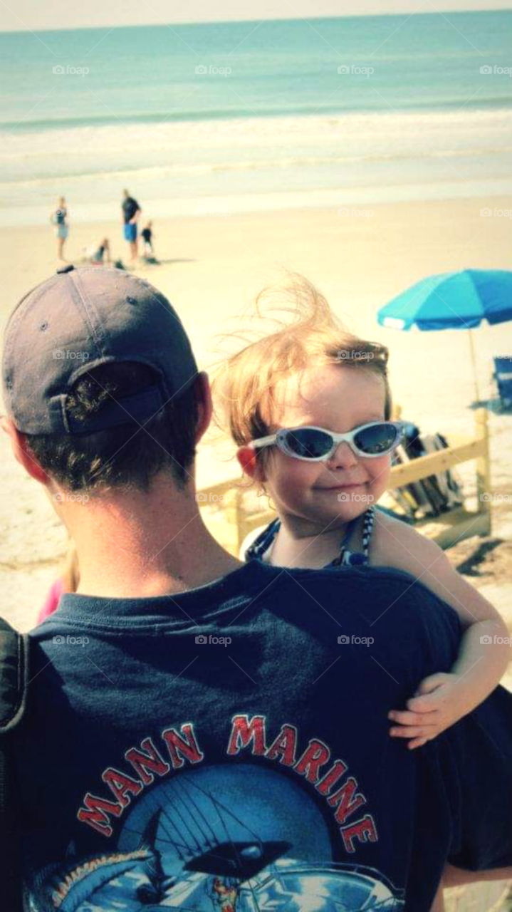 Father carrying young daughter who is looking over his shoulder wearing sunglasses to the beach