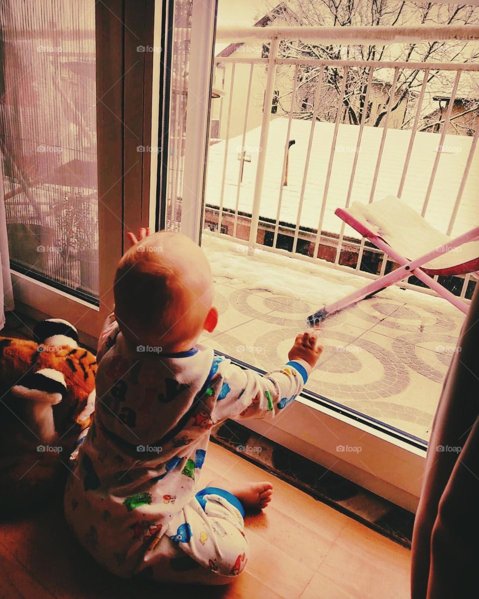 a one-year-old baby looks out the window while it is snowing