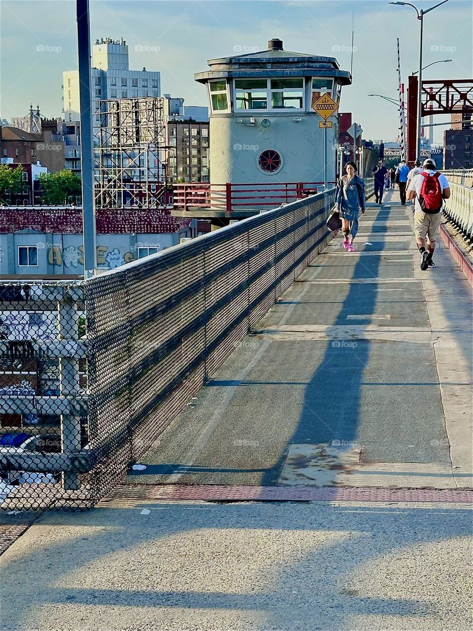 This is the “Pulaski Bridge” that connects “Greenpoint”, Bklyn to LIC, Queens in the direction we are headed as the sun casts dramatic shadows onto the pedestrian lane. The center outpost watchtower can be seen up ahead.. 2024. Hypnotic Productions