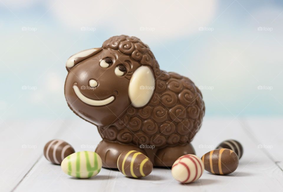 One cute chocolate Easter lamb with candy eggs around it stands on a white wooden table, vtd side close-up. Holiday treat concept, happy easter.