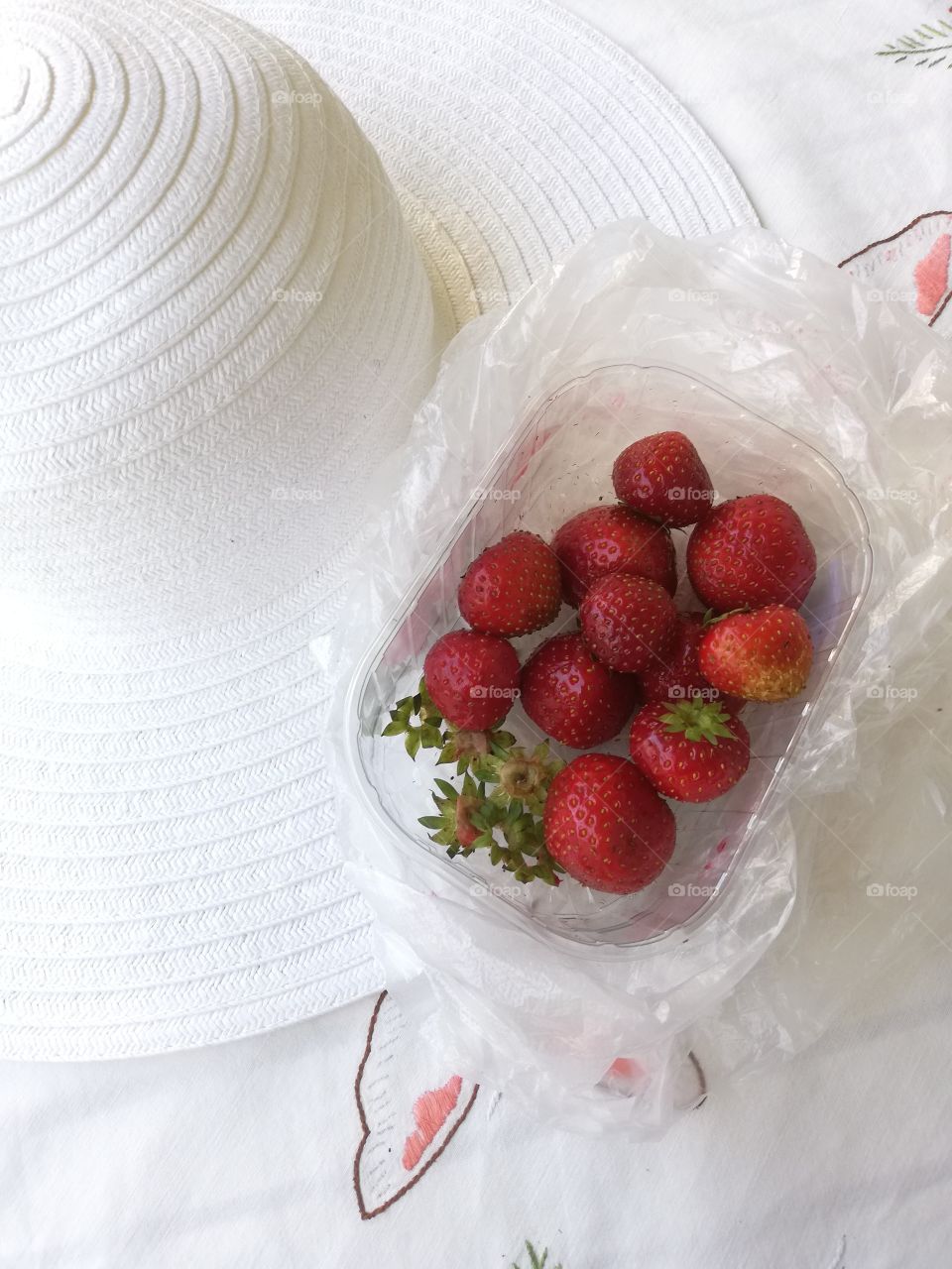 Bright red delicious fresh strawberries and some green leaves in a see-through not full plastic box are in a wrinkled plastic bag on an embroidered table cloth and on a brim of a white hat.