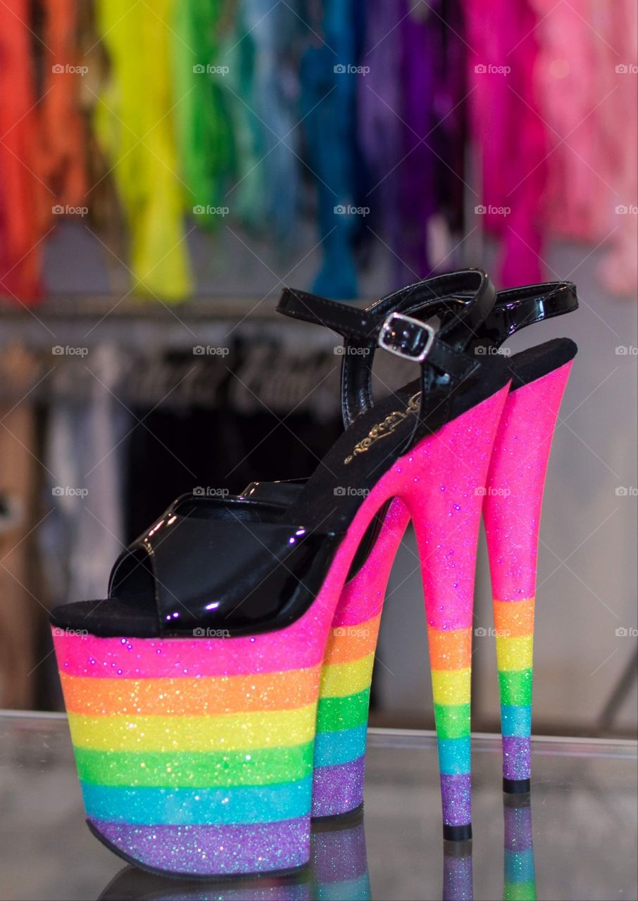 extremely bright rainbow colored high heel shoes in foreground with brightly colored bodysuits arranged in rainbow colors order and slightly blurred in background