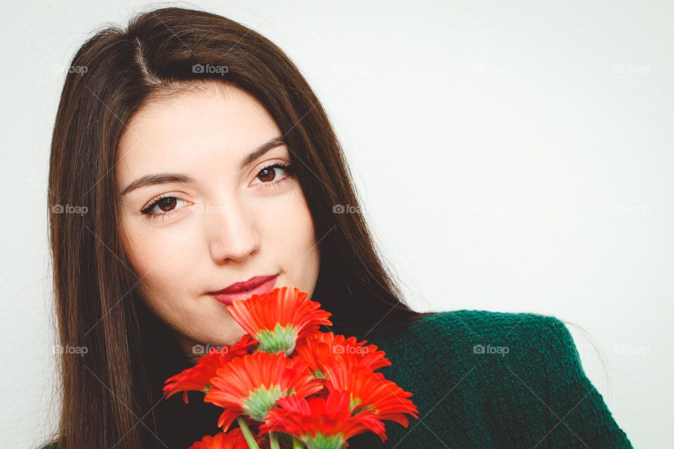 Portrait of a beautiful young Caucasian girl brunette with straight long hair holding a bouquet of red gerberas near her face and smiling at the camera against a gray-white background. The concept of beautiful people, healthy hair.