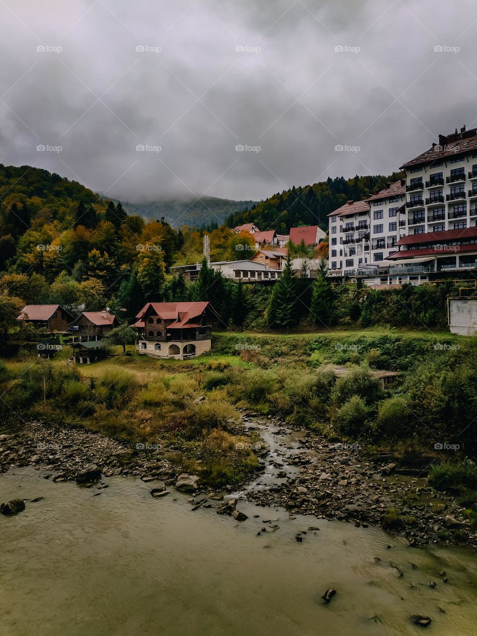 Small town in the mountains covered with forest in autumn. Red rooftops of multi storey buildings. Green grass and stones shore of the mountain river with dirty water. Living close to nature top view. Mountain tops in the fog, gray heavy clouds above