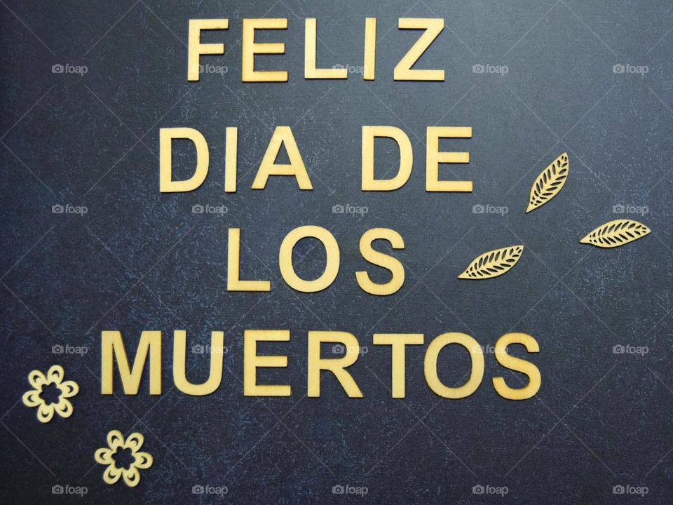 A simple seasonal phrase flatlay which reads “Feliz Dia De Los Muertos” spelled out in neutral wooden letters, accompanied by wooden flowers & leaves, laid out on a dark blue background.