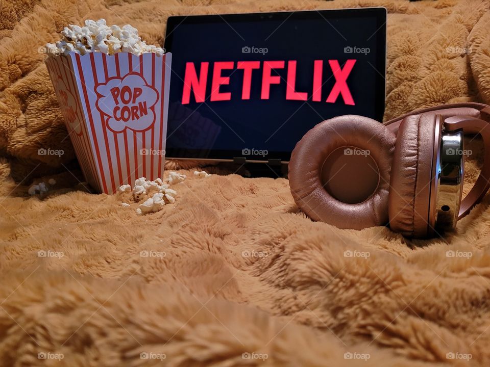 Laptop computer screen ready to watch Netflix for a fun, cozy, and relaxing night with popcorn as a snack, on soft bed blanket in room, with pink headphones