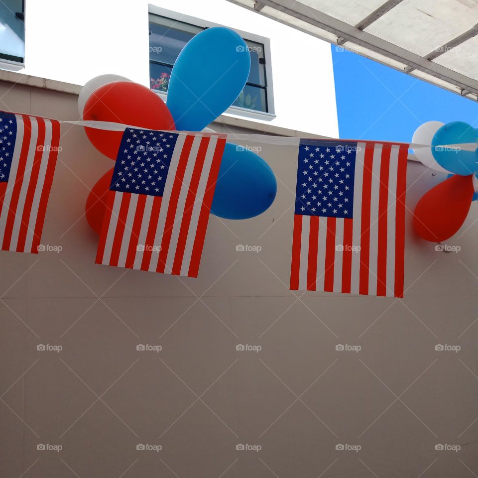 A string of American flags,  red,  white and blue baloons,  city backyard. A bit of blue sky,  roof