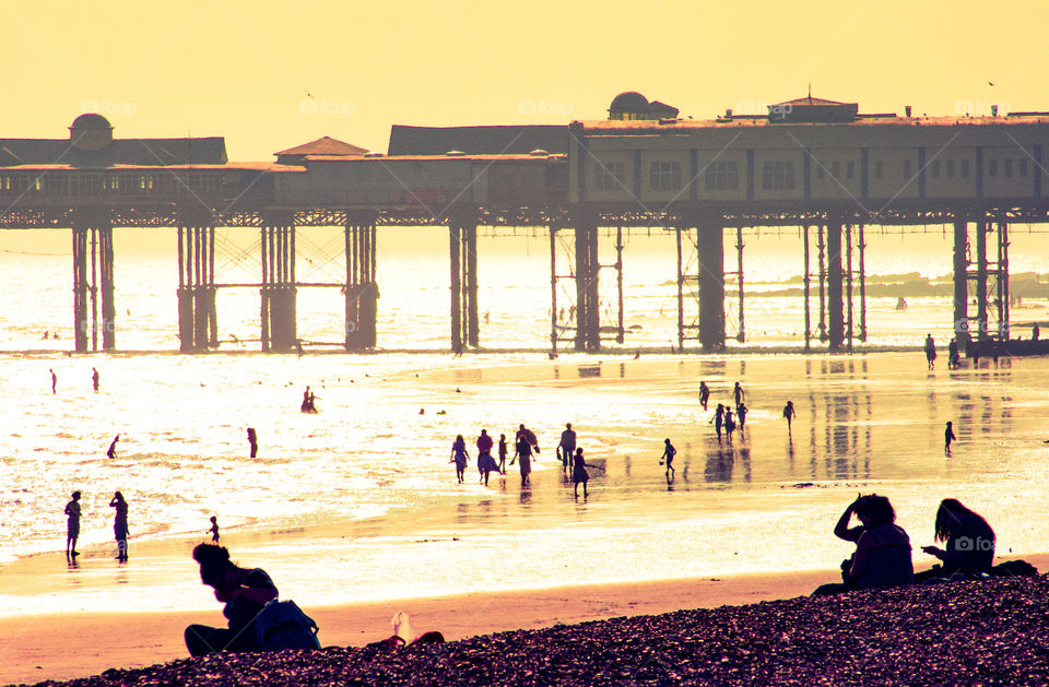 People are silhouetted against the yellow of the setting sun, on the beach. A pier is in the background 