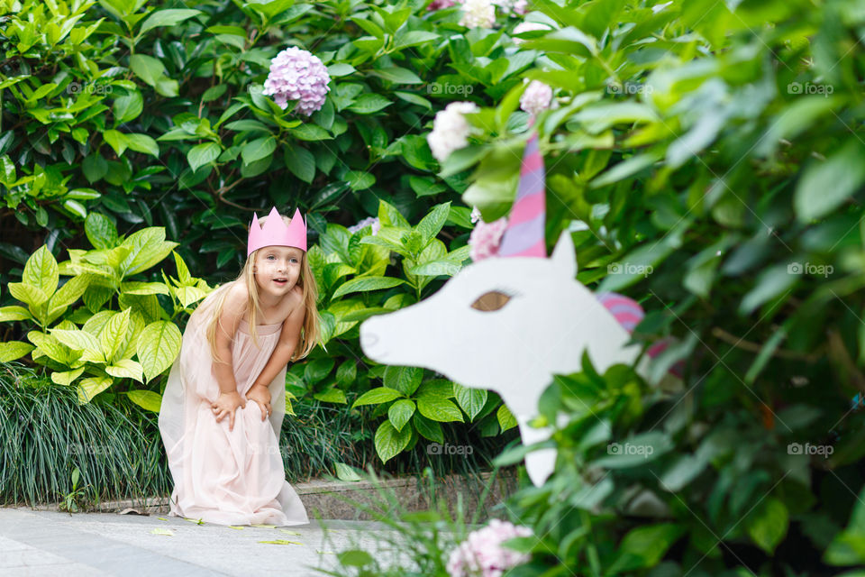 Happy little Caucasian girl with blonde hair and crown standing near unicorn in blooming garden at summer day