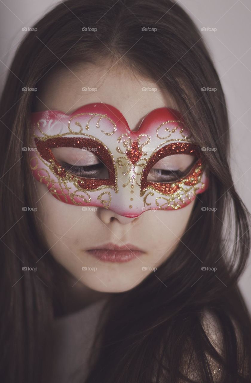 Portrait of a beautiful little Caucasian brunette girl with long flowing hair and a masquerade mask on her face looking down thoughtfully, side view close-up. The concept of carnival, masquerade.