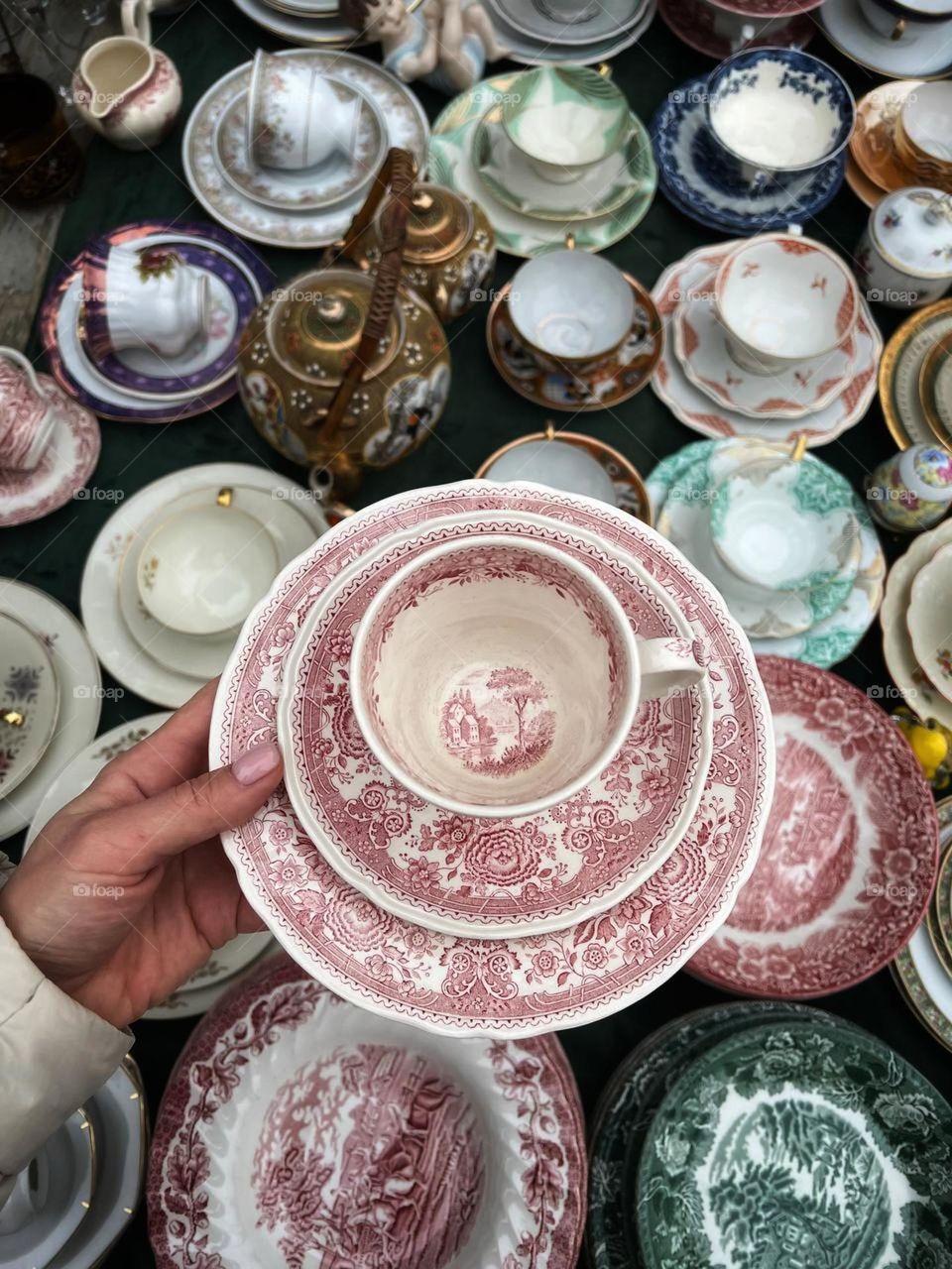 Vintage collectible porcelain tableware spread out on a table. A woman holds in her hand a tea pair with a red and white pattern. A variety of cups, saucers and plates with different patterns and colors are arranged around them