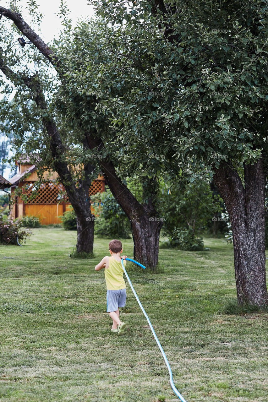 Boy pulling a garden hose to watering the plants in backyard garden. Candid people, real moments, authentic situations