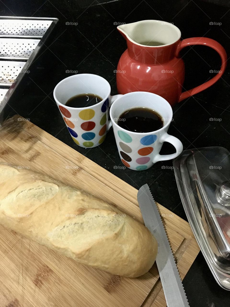 Freshly home-made French bread sitting on a bamboo cutting board next to two mugs of tea, a red pitcher, a butter dish and a bread knife.