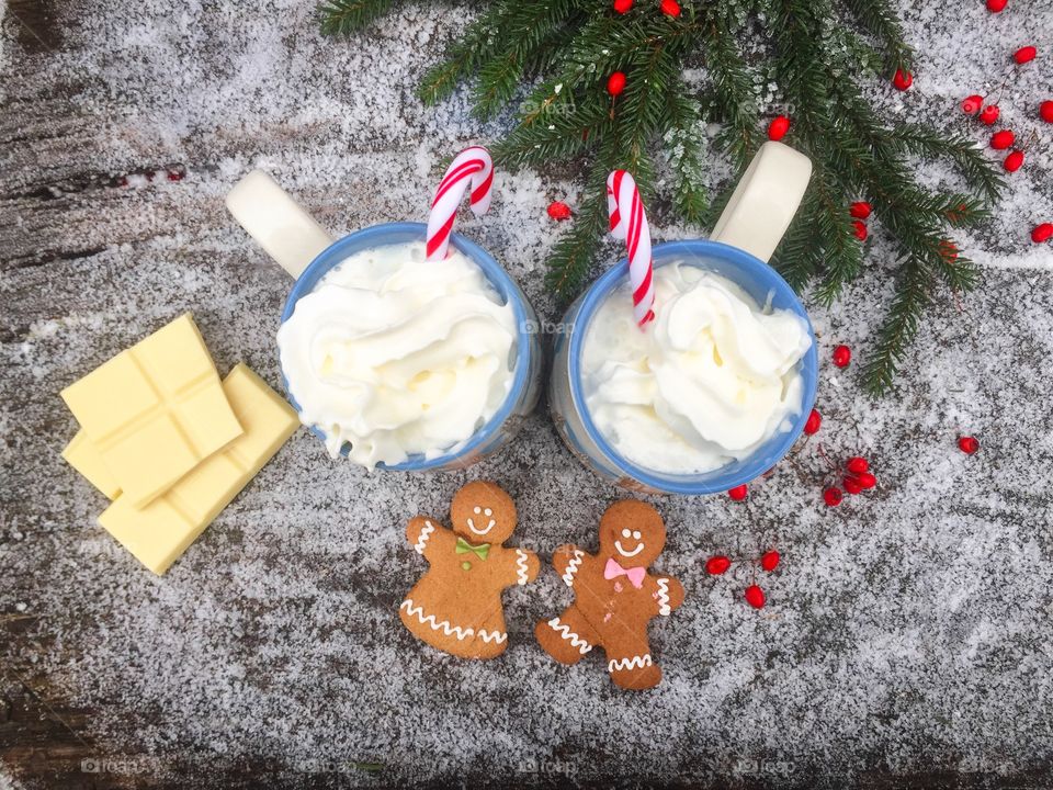 Two mugs of white chocolate with whipped cream, gingerbread man and candy canes on a snowy table with pieces of white chocolate and evergreen branches beside