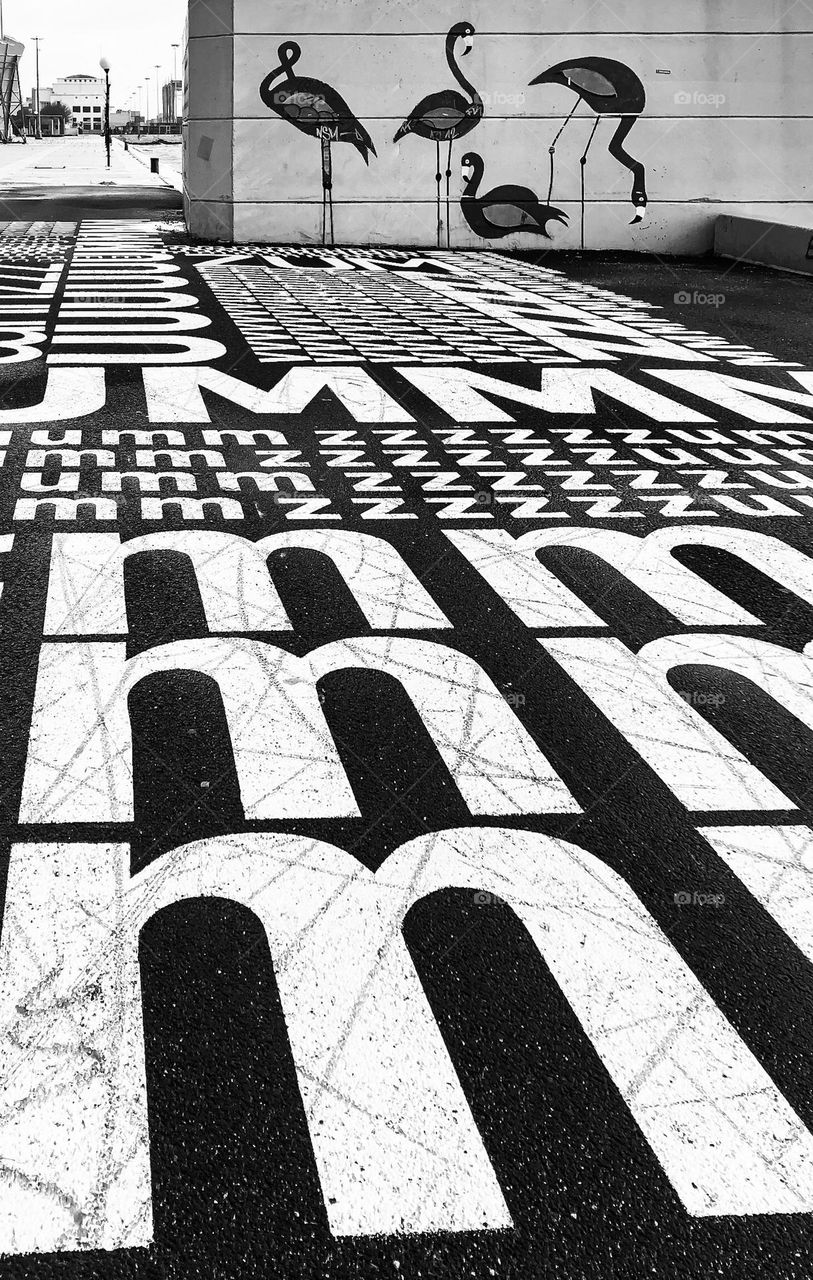 Black and white image of letter painted on the ground and flamingo like birds on the wall below Ponte 25 de Abril, Lisboa