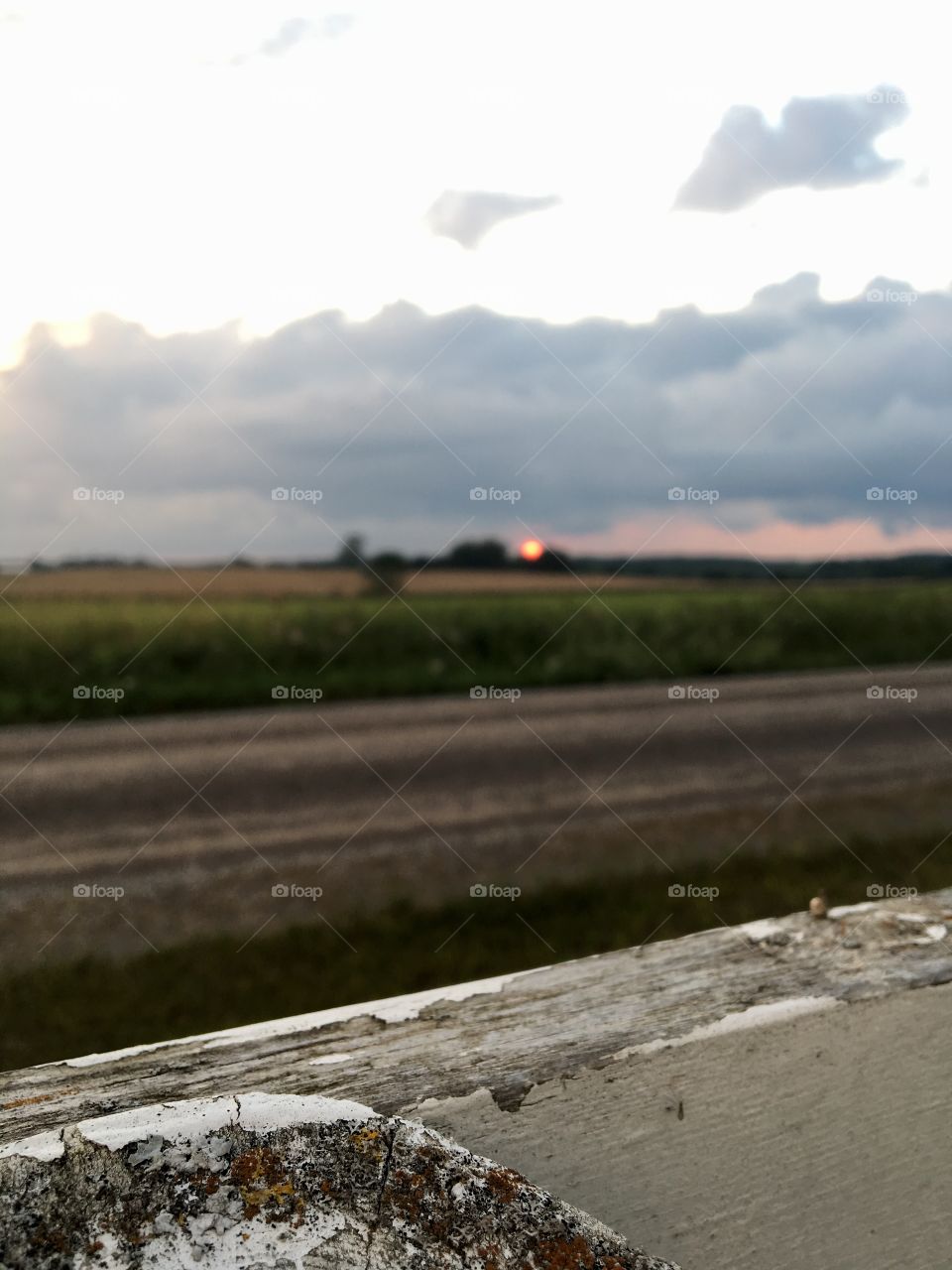 Fluffy forming storm clouds overtop a setting sun. In the foreground is a aged white fence. Behind the fence is a gravel road, then endless fields.