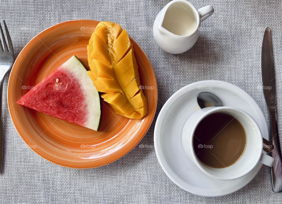 One slice of watermelon and mango on an orange round plate. Coffee and milk on the gray table for breakfast. 