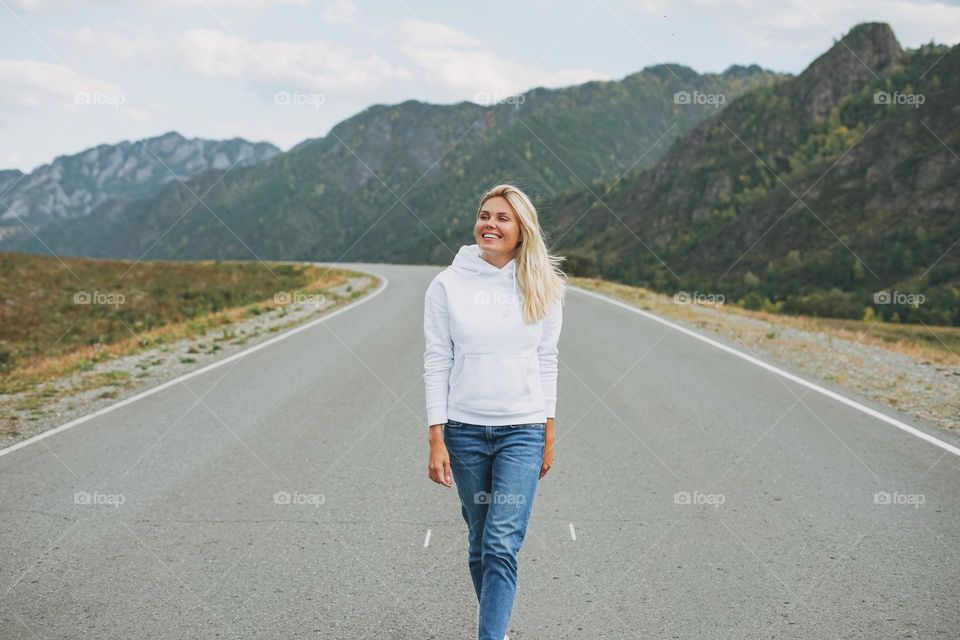 Beautiful smiling blonde young woman traveler in white hoodie on road, trip to mountains