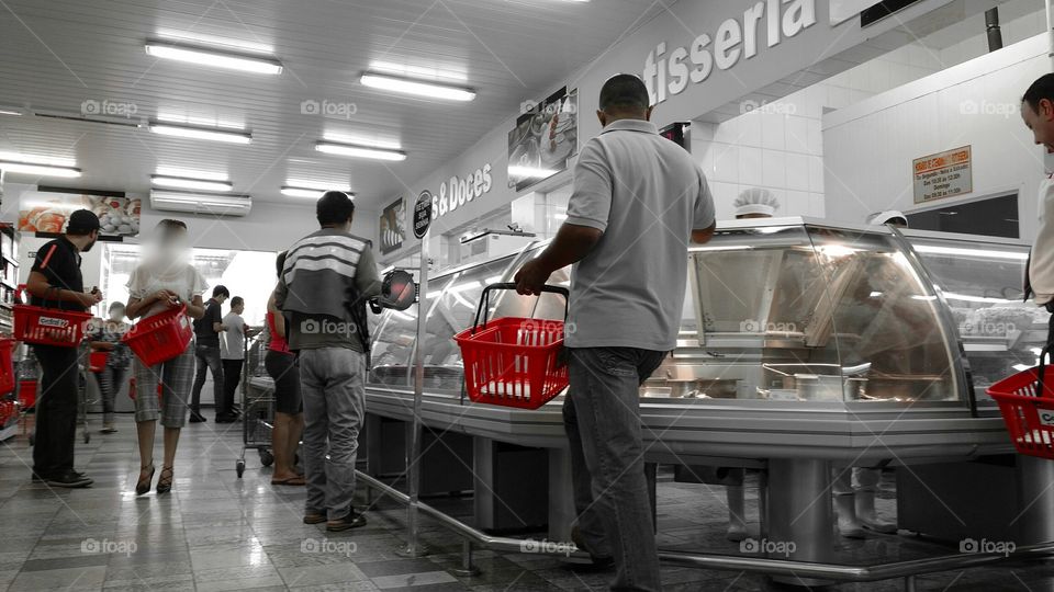 Pessoas com cestinhas plásticas, aguardando chamada de senhas em Rotisseria e Restaurante, dentro de Supermercado.