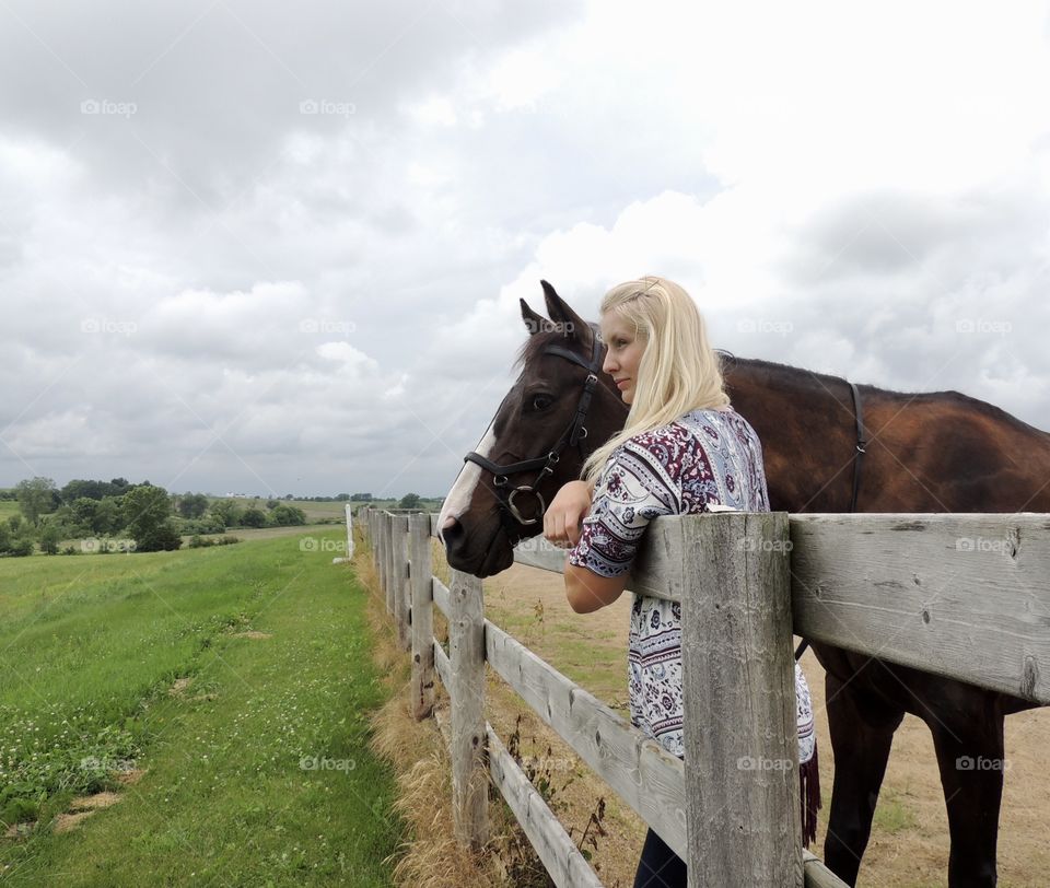 Blonde girl and dark bay horse standing side-by-side looking over a wooden fence separating the arena and pasture.