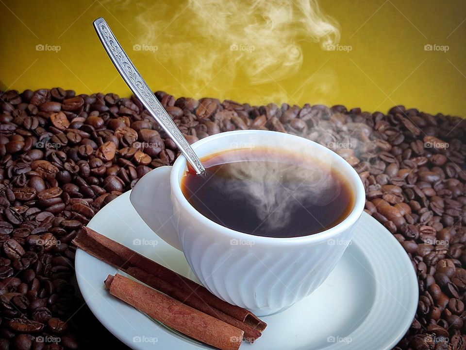 A white cup of black coffee stands on a white sauce with two cinnamon sticks. There is a metal spoon in the cup. Steam comes out of a cup of coffee. The composition stand on coffee beans. In the background is a yellow background